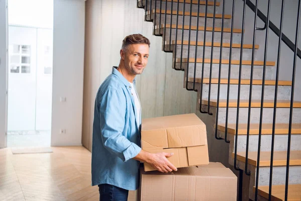Bonito homem segurando caixa em mãos e sorrindo perto de escadas em nova casa — Fotografia de Stock