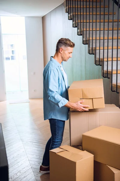 Handsome man looking at box in hands while standing near stairs in new home — Stock Photo