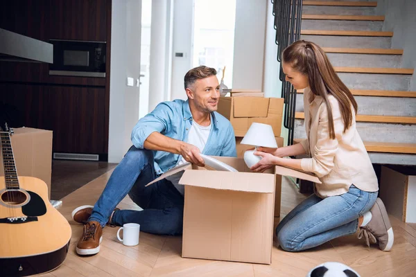 Alegre marido mirando esposa desembalaje caja y sosteniendo lámpara en las manos mientras está sentado en el suelo - foto de stock