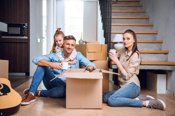 Cheerful kid hugging dad sitting on floor with cup of drink near wife — Stock Photo