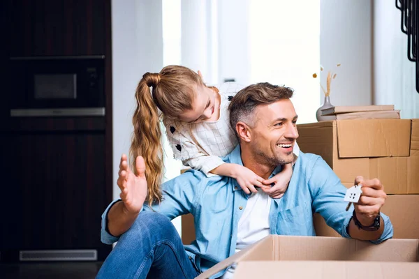 Cheerful child hugging smiling father sitting on floor with house shaped key chain — Stock Photo