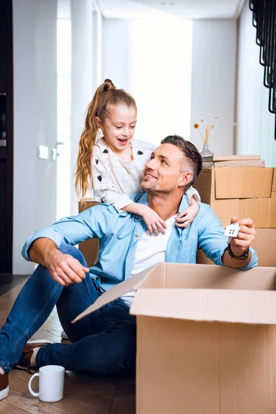 Cheerful daughter looking at father with house shaped key chain sitting on floor — Stock Photo