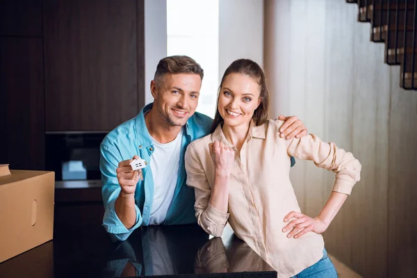 Bonito homem abraçando esposa e segurando casa em forma de chaveiro na mão — Fotografia de Stock