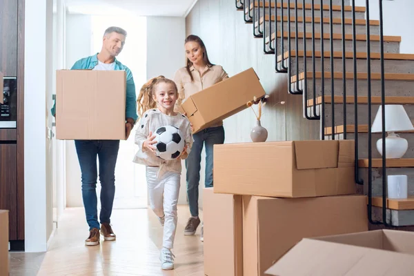 Niño alegre sosteniendo el fútbol con padres felices con cajas en el fondo - foto de stock