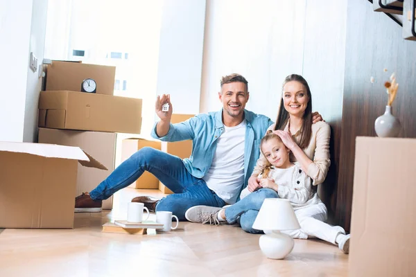 Happy man sitting on floor with house shaped key chain near attractive wife and daughter — Stock Photo