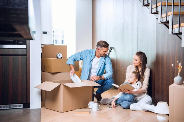 Woman holding book near daughter while sitting on floor and looking at husband — Stock Photo