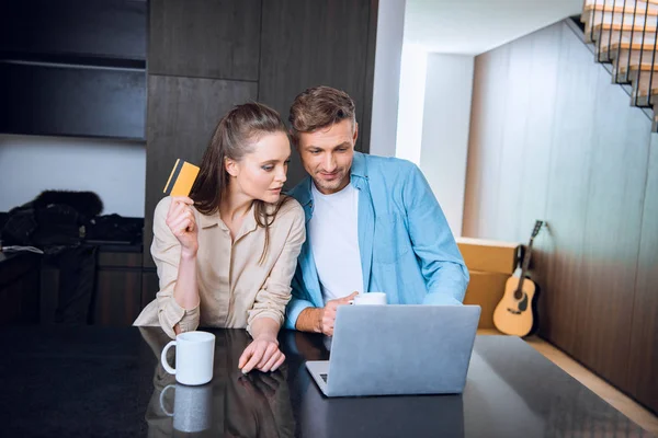 Hombre feliz mirando el ordenador portátil cerca atractiva esposa celebración de la tarjeta de crédito - foto de stock