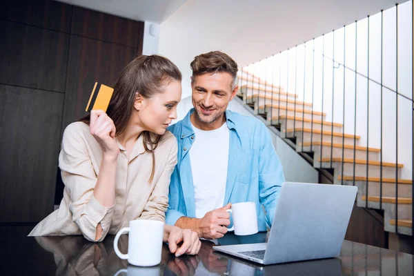 Homem feliz olhando atraente esposa segurando cartão de crédito perto do laptop — Fotografia de Stock