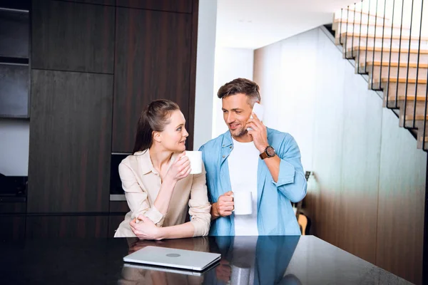 Fröhliche Frau hält Tasse Kaffee in der Hand und schaut Ehemann beim Smartphone-Gespräch an — Stockfoto