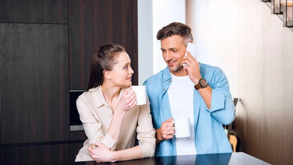 Attractive woman holding cup of coffee and looking at husband talking on smartphone — Stock Photo
