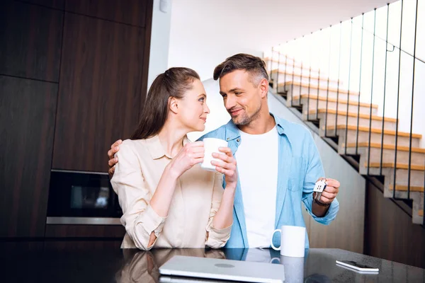 Mulher atraente com xícara de café olhando para marido alegre segurando casa em forma de chaveiro — Fotografia de Stock