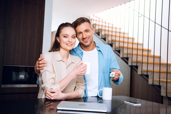 Mulher atraente segurando xícara de café e de pé com marido alegre segurando casa em forma de chaveiro — Fotografia de Stock