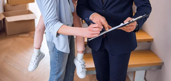 Cropped view of woman holding in arms daughter and signing document broker holding clipboard — Stock Photo