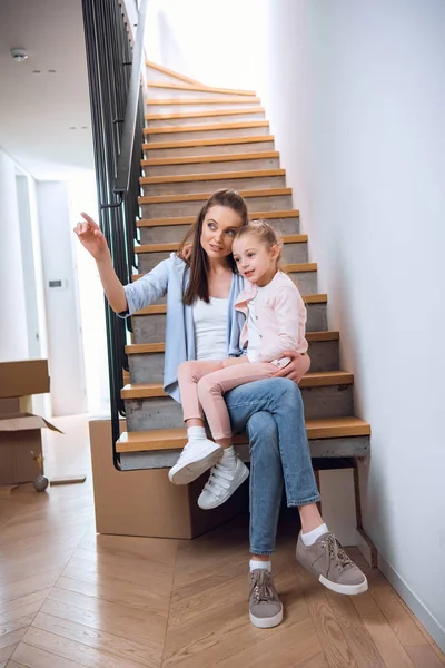 Attrayant femme assise avec fille dans les escaliers et pointant du doigt dans la nouvelle maison — Photo de stock