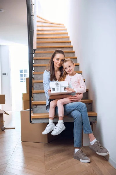 Mujer feliz sentada con su hija en las escaleras y sosteniendo modelo de casa - foto de stock