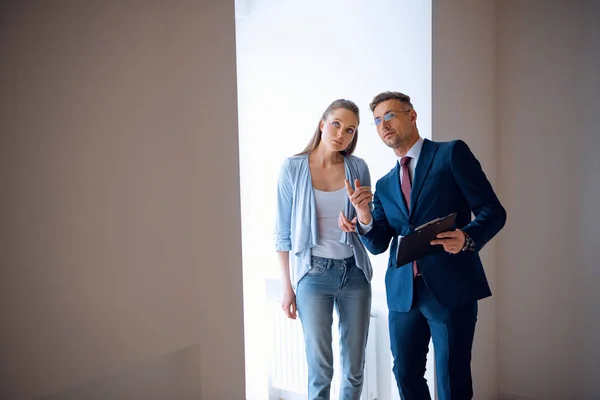 Schöner Makler, der mit dem Finger zeigt, während er mit attraktiver Frau im Zimmer steht — Stockfoto