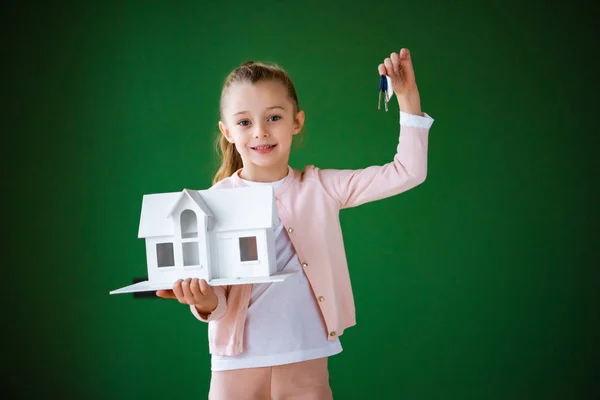 Niedliches Kind hält Hausmodell und Schlüssel in den Händen auf grünem Hintergrund — Stockfoto