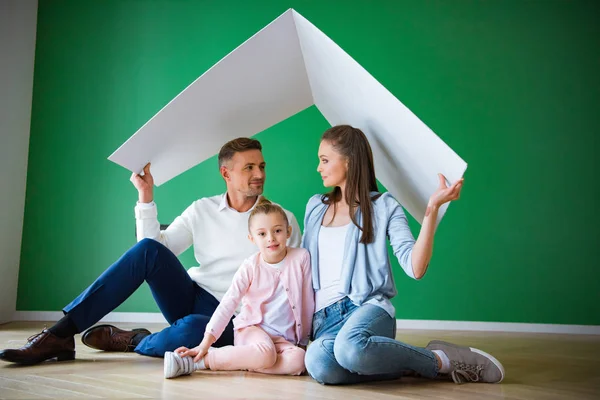 Happy husband and wife looking at each other near cute daughter and holding paper roof while sitting on floor — Stock Photo