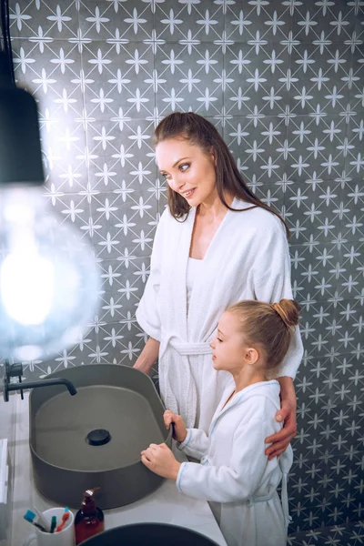 Hermosa mujer de pie con linda hija en batas de baño - foto de stock