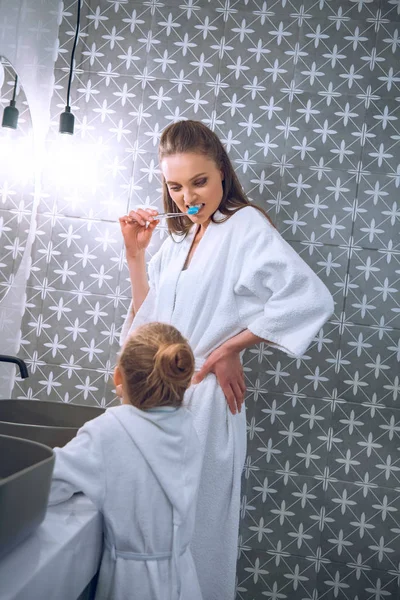 Atractiva mujer cepillarse los dientes cerca de hija en bata de baño - foto de stock