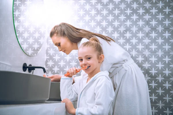 Lindo niño cepillarse los dientes cerca de la madre en bata de baño - foto de stock