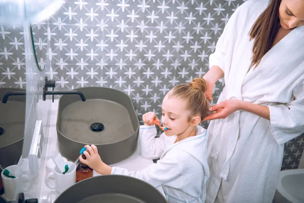 Adorable enfant brossant les dents près de mère en peignoir de bain — Photo de stock