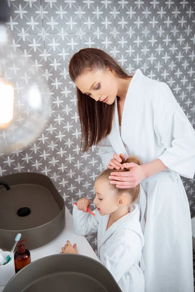 Mignon enfant brossant les dents près de belle mère en peignoir de bain — Photo de stock