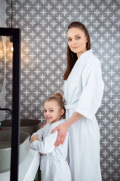 Feliz madre de pie con hija en batas de baño - foto de stock