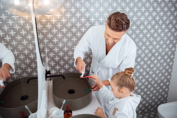 Mann legt Zahnpasta auf Zahnbürste von Tochter im Bademantel — Stockfoto