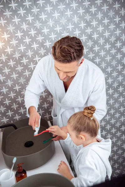 Dad putting toothpaste on toothbrush of daughter in bathrobe — Stock Photo