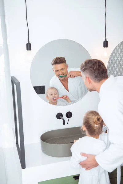 Усміхнений батько і дочка чистять зуби в банних халатах вдома — стокове фото