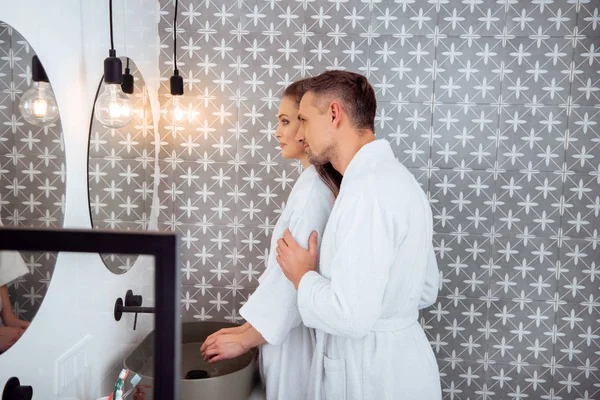 Husband hugging attractive wife in bathroom while standing in white bath robe — Stock Photo