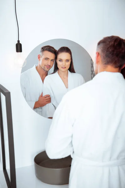 Красивий чоловік обіймає дружину у ванній, стоячи в білому халаті і дивлячись на дзеркало — стокове фото