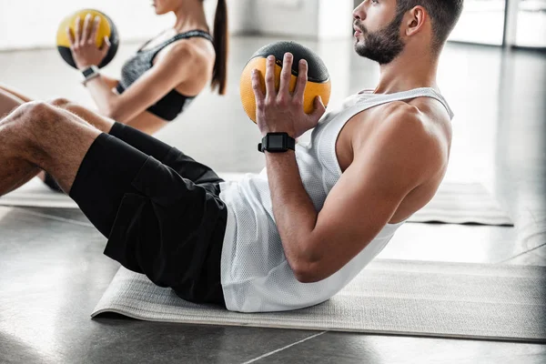 Recortado disparo de atlético joven pareja sosteniendo bolas de medicina y haciendo abdominales ejercicio en esteras de yoga - foto de stock