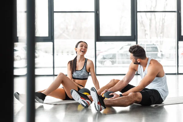 Alegre pareja joven deportiva sentada en colchonetas de yoga con bolas de medicina y sonriéndose mutuamente en el gimnasio - foto de stock