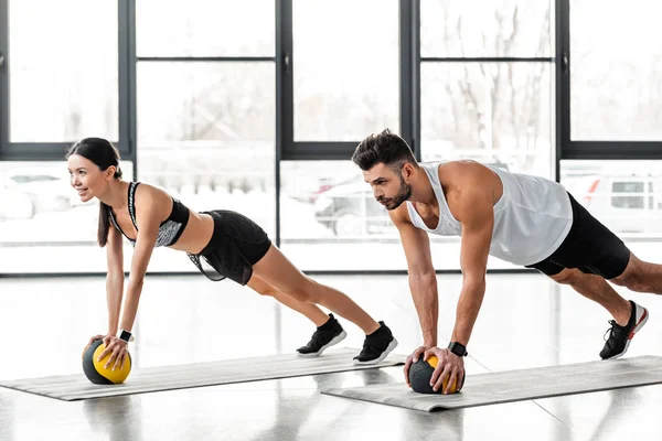 Jovem casal atlético em sportswear exercício com bolas de medicina em tapetes de ioga e olhando para longe no ginásio — Fotografia de Stock