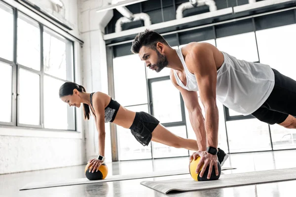Vista lateral de pareja joven atlética en ropa deportiva haciendo ejercicio con pelotas de medicina en colchonetas de yoga en el gimnasio - foto de stock