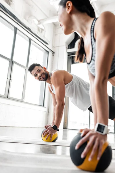Pareja joven atlética sonriéndose mientras hacen ejercicio con pelotas de medicina en colchonetas de yoga en el gimnasio - foto de stock