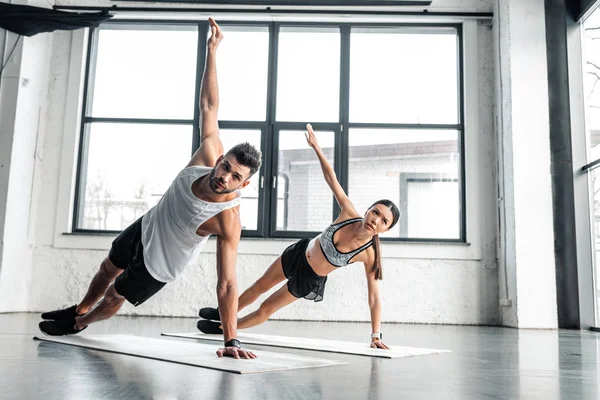 Vista completa de la pareja joven atlética haciendo ejercicio de tablón lateral en esteras de yoga en el gimnasio - foto de stock