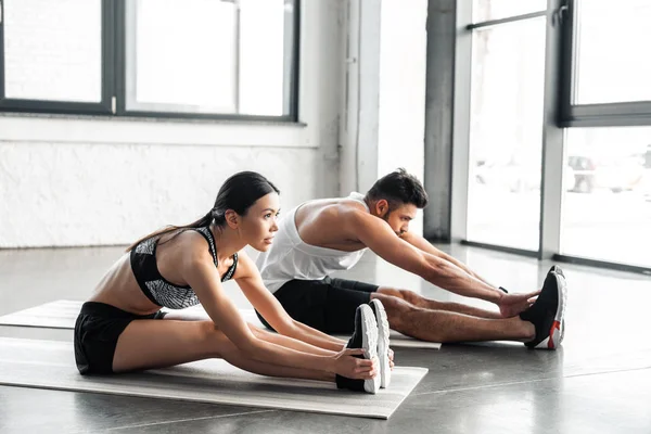 Вид сбоку спортивной молодой пары, растягивающей и упражняющейся на ковриках для йоги в тренажерном зале — стоковое фото