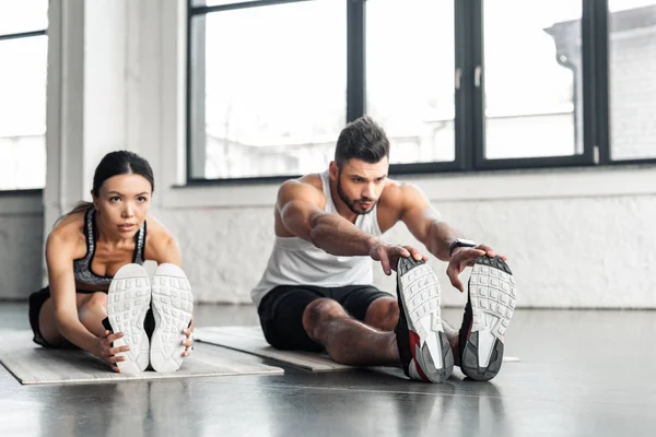 Pareja joven y deportiva estirando las piernas y haciendo ejercicio en colchonetas de yoga en el gimnasio - foto de stock