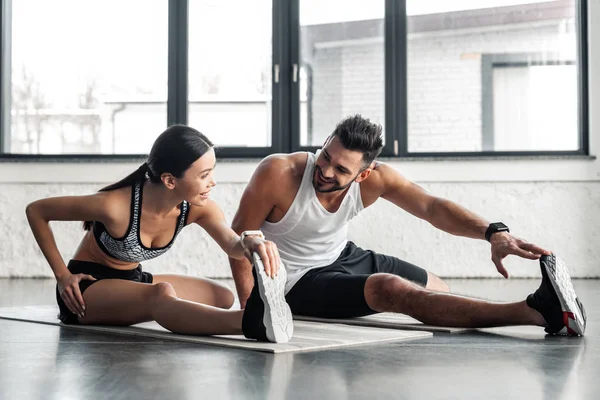 Pareja joven y deportiva estirando las piernas y sonriéndose mientras entrenan en colchonetas de yoga en el gimnasio - foto de stock