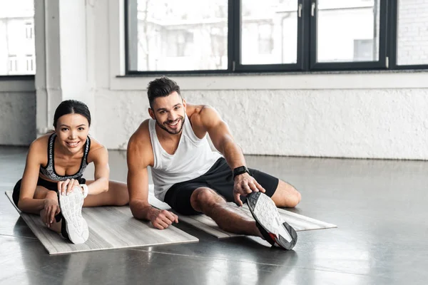 Cheerful young sporty couple stretching on yoga mats and smiling at camera in gym — Stock Photo