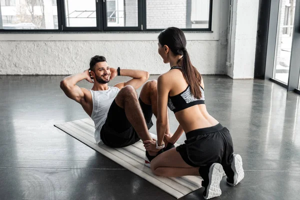Chica deportiva ayudando a hombre joven feliz haciendo ejercicios abdominales en la estera de yoga en el gimnasio - foto de stock