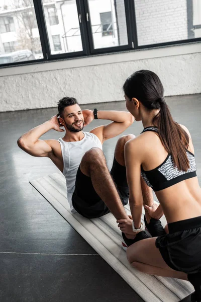 Vista de ángulo alto de chica deportiva ayudando a hombre joven feliz haciendo ejercicios abdominales en estera de yoga en el gimnasio - foto de stock
