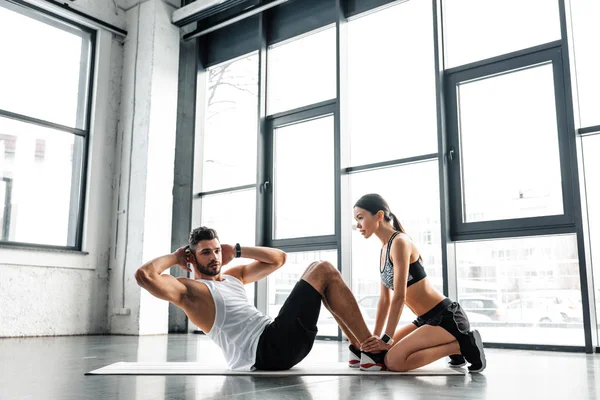 Side view of young sportswoman holding legs of athletic man doing abs exercise in gym — Stock Photo