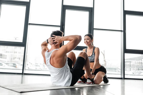 Улыбающаяся спортивная девушка помогает юному спортсмену делать упражнения на прессе на коврике для йоги в спортзале — стоковое фото