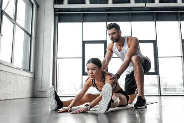 Bonito atlético homem ajudando desportivo mulher alongamento no ioga tapete no ginásio — Fotografia de Stock