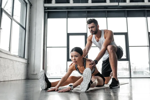 Bell'uomo atletico che aiuta la donna sportiva che si allunga sul tappetino di yoga in palestra — Foto stock