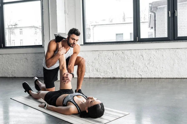 Entrenador masculino ayudando a la joven deportista a estirar la pierna en la esterilla de yoga en el gimnasio - foto de stock